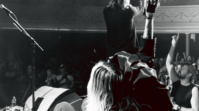 Black and white image of the members of 'Band of Skulls' on stage performing taking from behind them and the crowd is in front with a man in the bottom right with his hand raised in the air.
